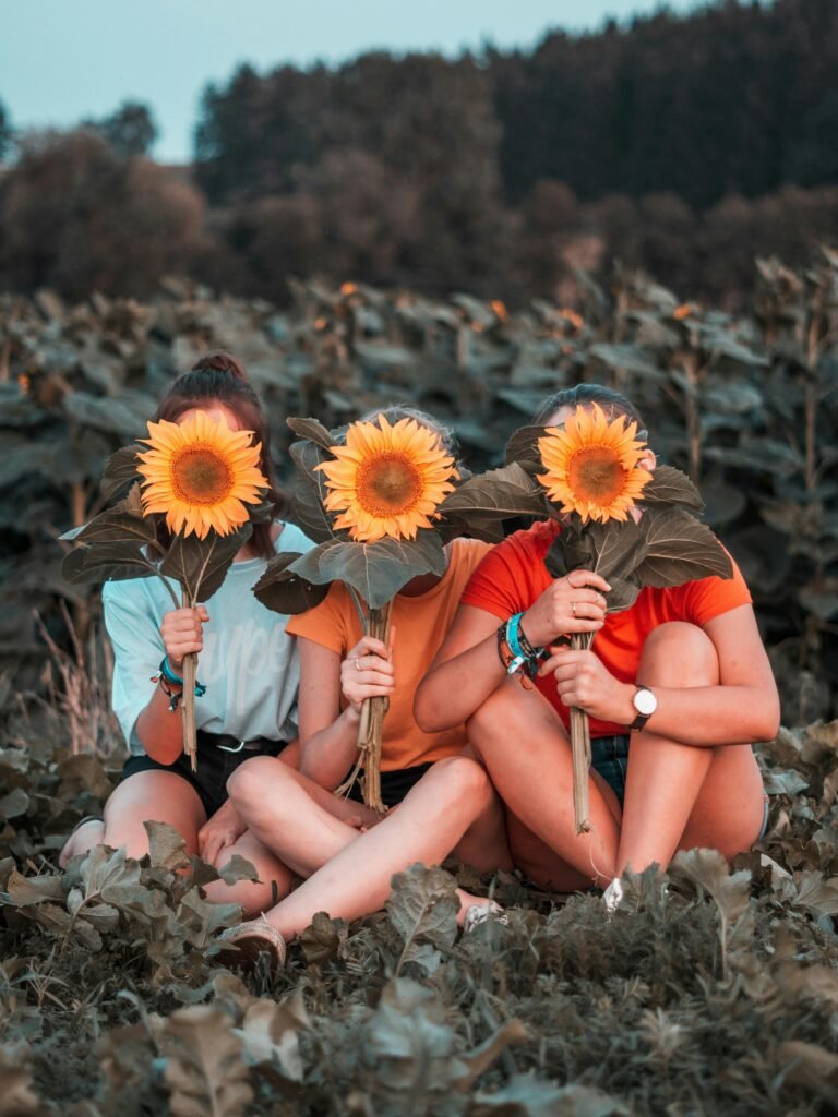 Three women sitting in a sunflower field in Austria, playfully covering faces with sunflowers.